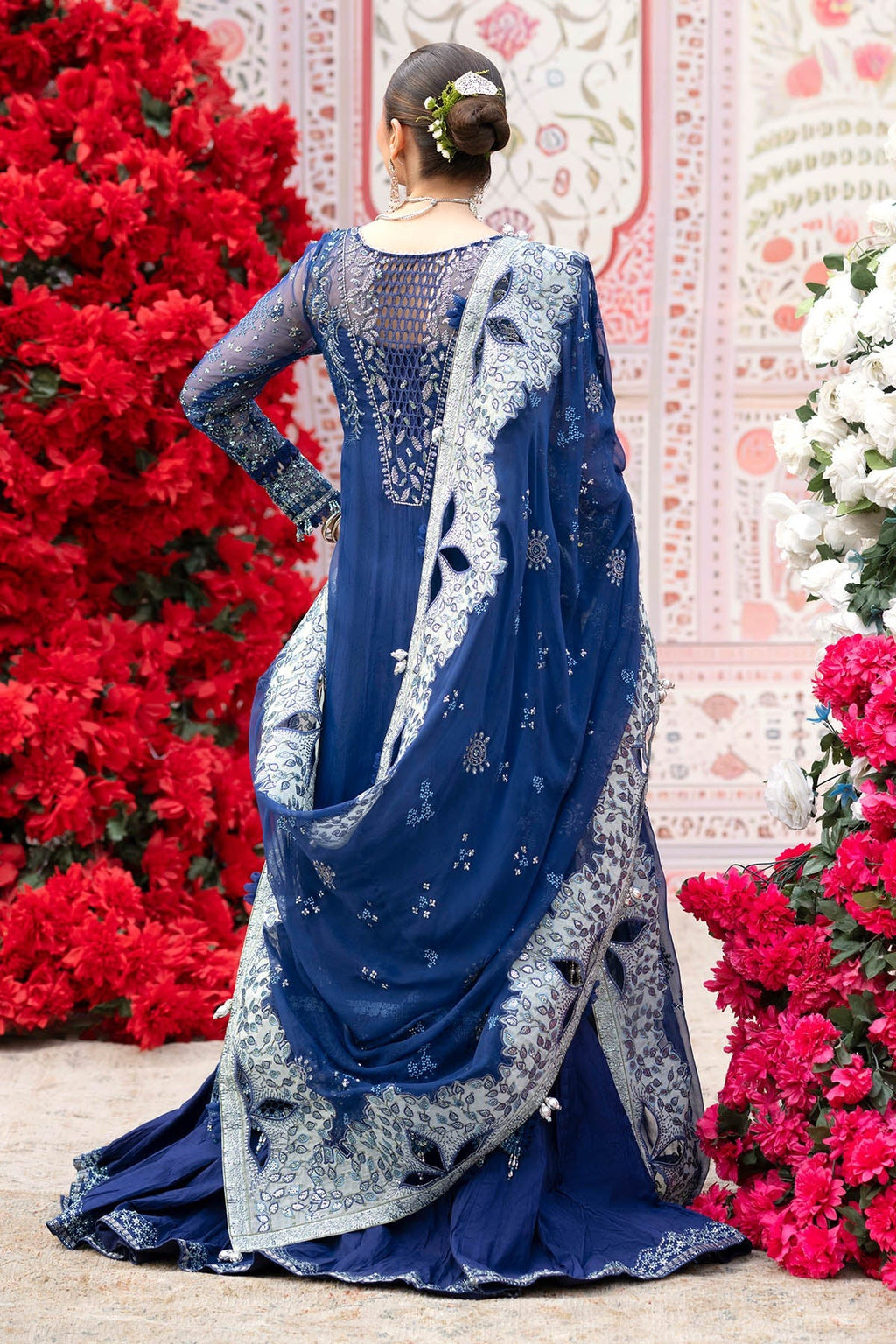 A woman wearing a royal blue embroidered traditional outfit with a matching sheer dupatta, photographed from the back in front of a decorative pink wall and red and white floral arrangements.