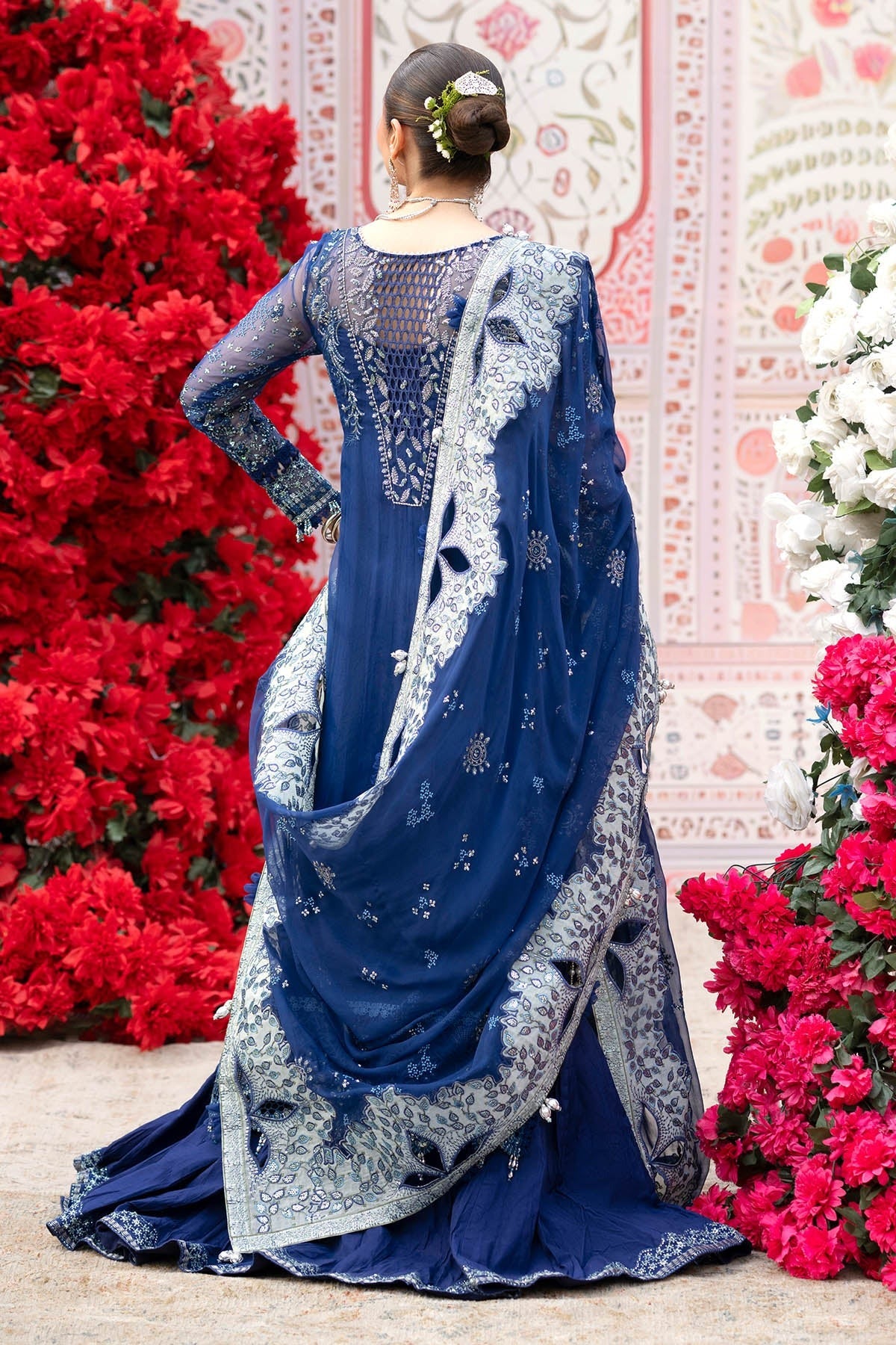 A woman wearing a royal blue embroidered traditional outfit with a matching sheer dupatta, photographed from the back in front of a decorative pink wall and red and white floral arrangements.