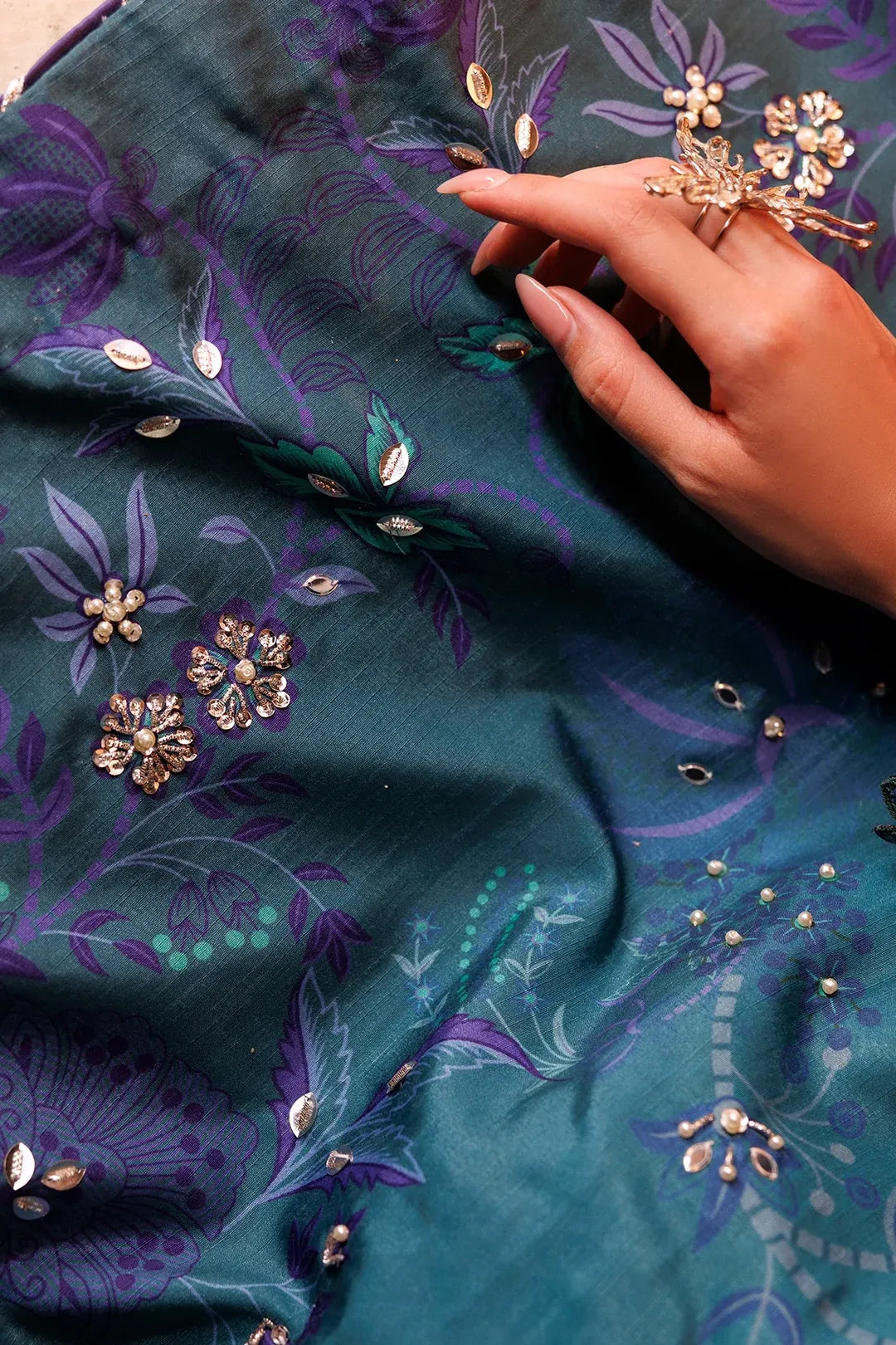 A close-up of a teal green fabric with purple floral patterns and gold mirror work embellishments, held by a hand wearing a matching gold ring.