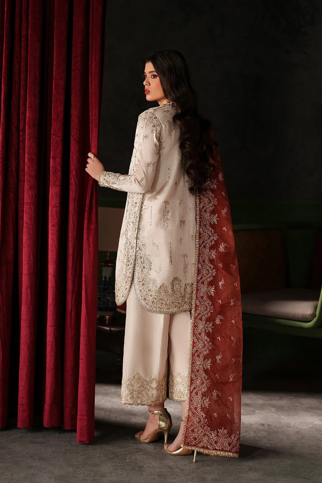 A woman stands in profile wearing a traditional South Asian outfit with a cream embroidered kurta and wide-leg pants, paired with a rust-red dupatta featuring gold floral embroidery. She is posed in front of a dark red velvet curtain in a dimly lit room.