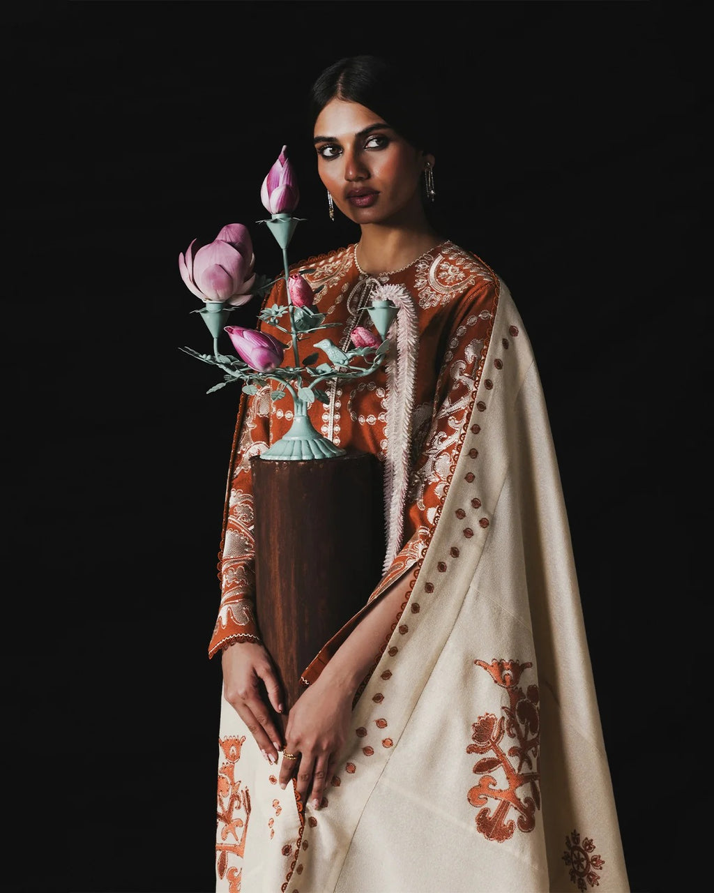 A woman models a traditional South Asian outfit featuring a cream-colored sari with rust-orange floral embroidery and a matching embroidered kurta. She holds a decorative vase with pink lotus flowers against a solid black background.