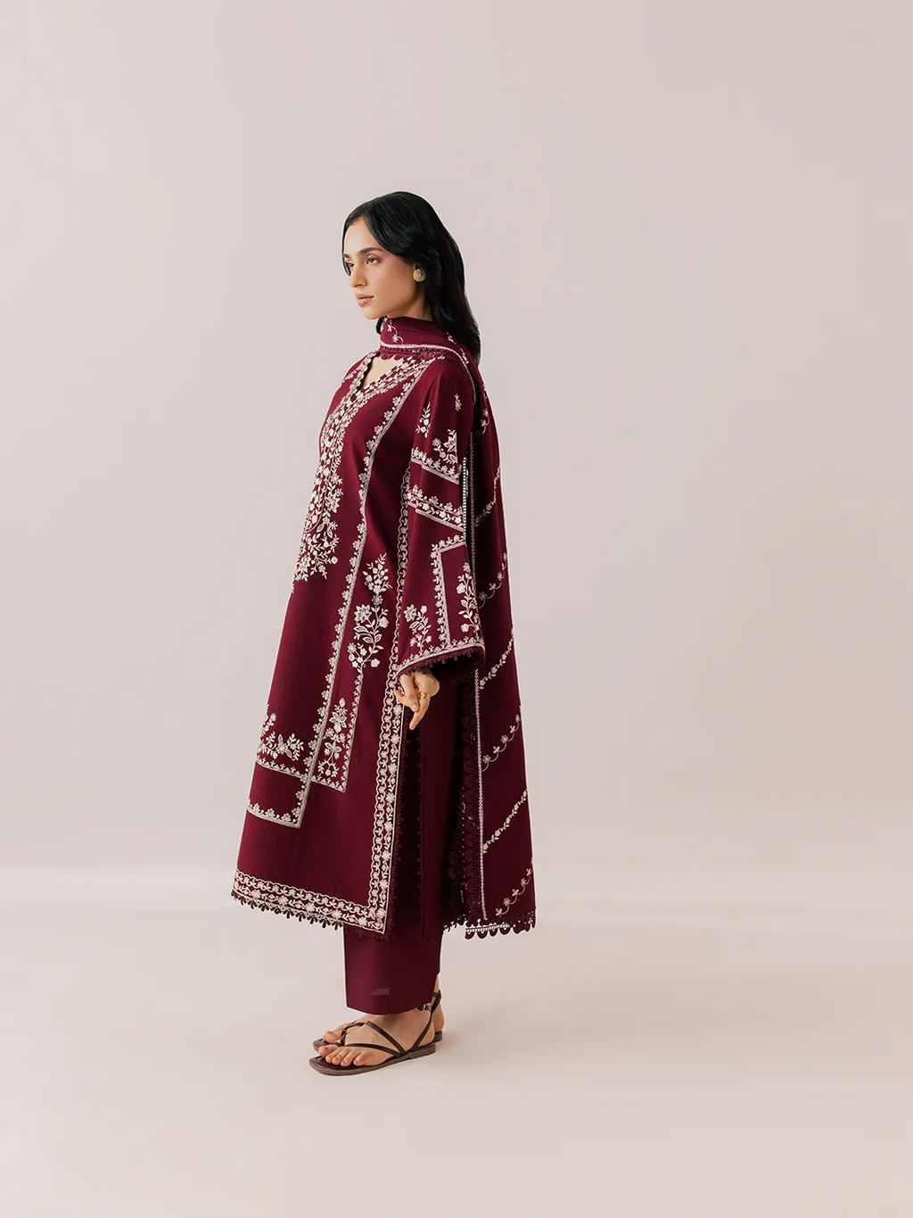 A woman stands in profile wearing a long maroon kaftan-style tunic with white geometric embroidery, matching maroon pants, and brown strappy sandals against a plain light beige studio background.