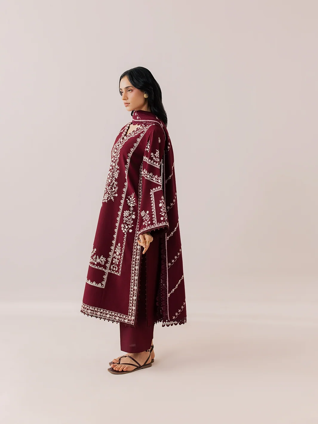 A woman stands in profile wearing a long maroon kaftan-style tunic with white geometric embroidery, matching maroon pants, and brown strappy sandals against a plain light beige studio background.