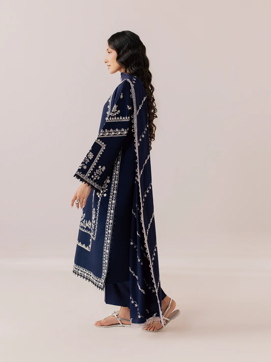 A woman modeling a navy blue traditional outfit with intricate white embroidery, shown from a side profile against a solid pale pink studio background.