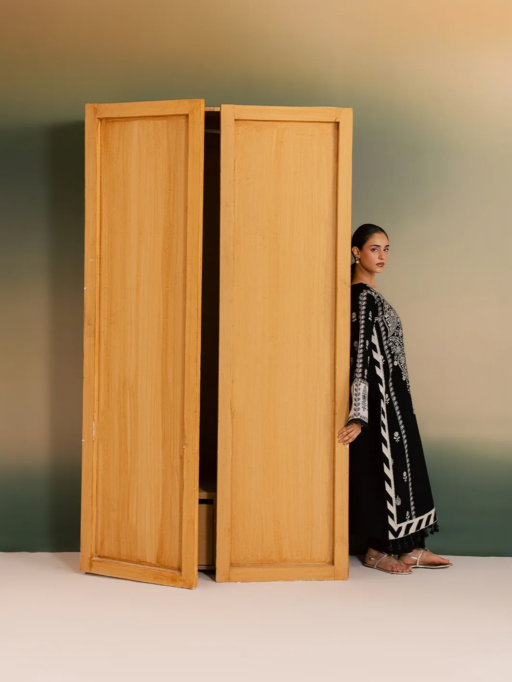 A woman wearing a black and white patterned traditional outfit stands partially behind two tall light wood doors against a gradient studio backdrop.