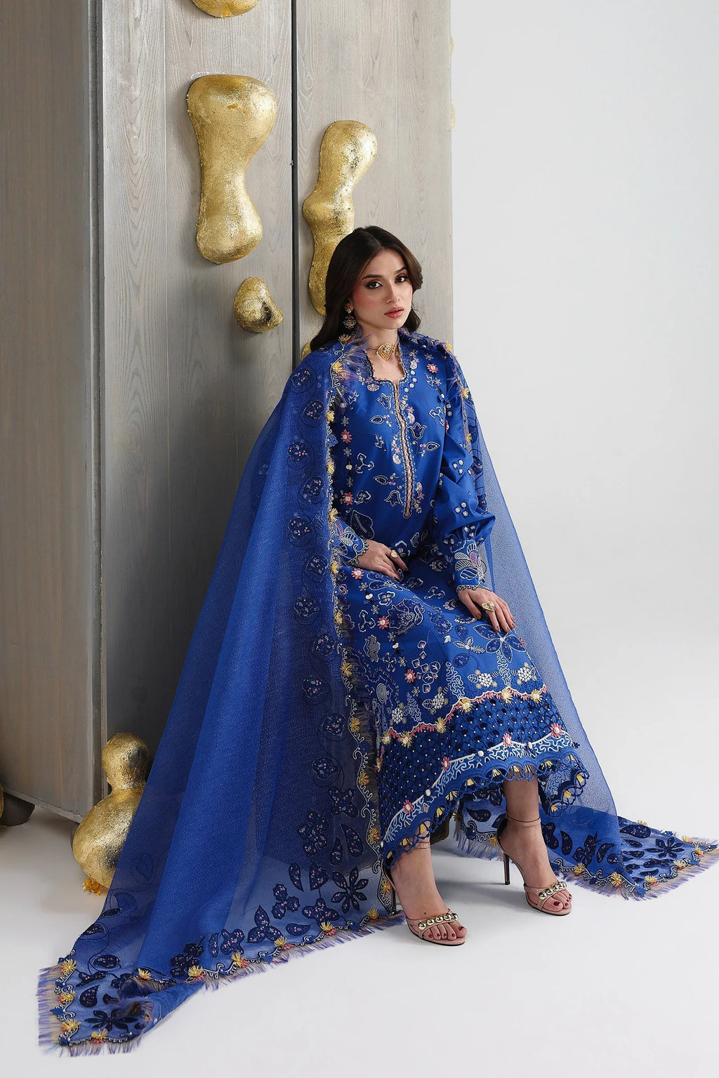 Woman in a blue embroidered traditional outfit sitting against a white wall with decorative elements.
