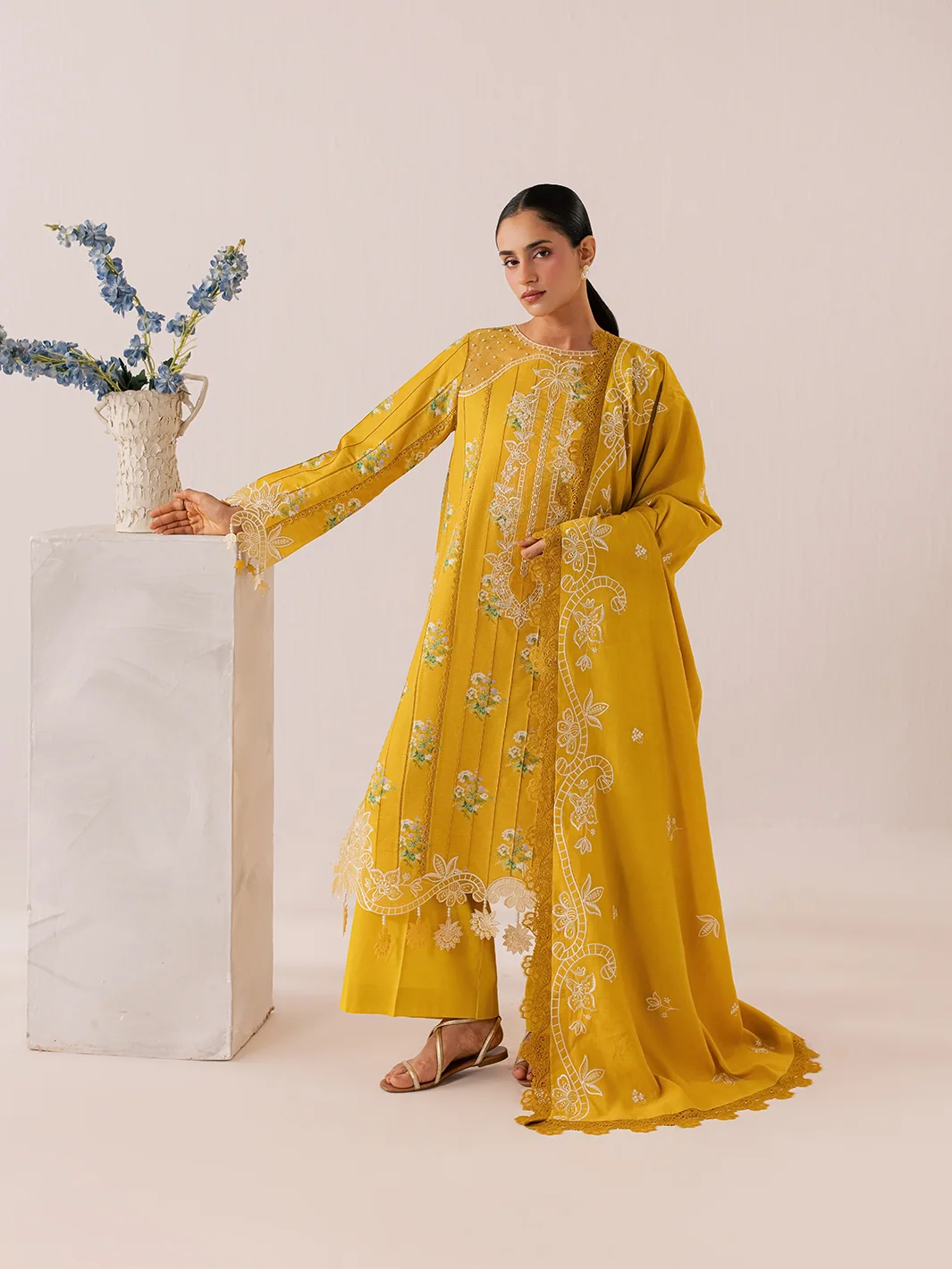 A woman models a traditional long yellow kurta set with intricate white floral embroidery and lace borders, standing next to a white pedestal with a vase of blue flowers against a solid beige background.