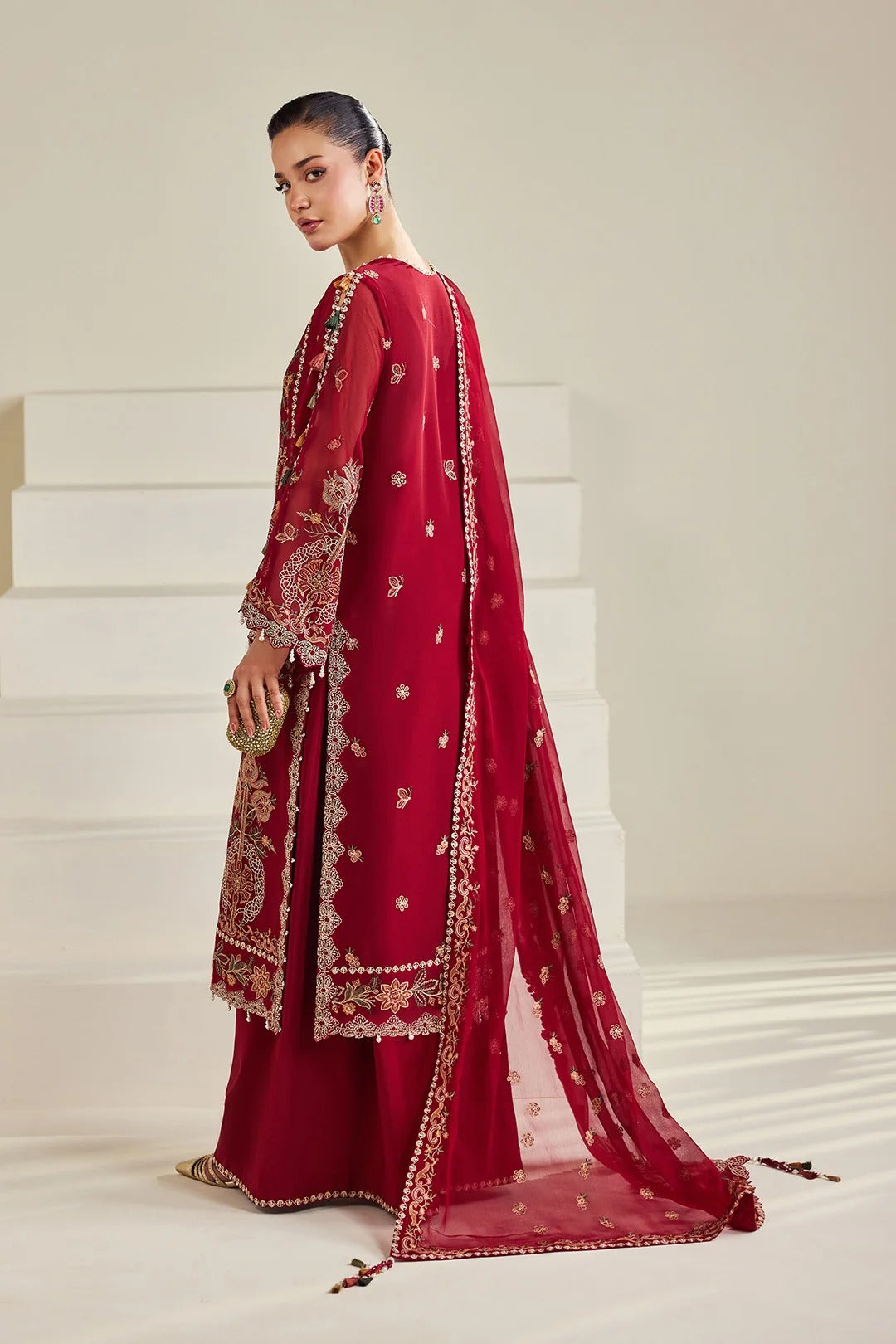 A woman models a traditional red Indian ethnic outfit with a matching sheer dupatta, featuring intricate gold embroidery and border details, set against a minimalist beige background with white steps.