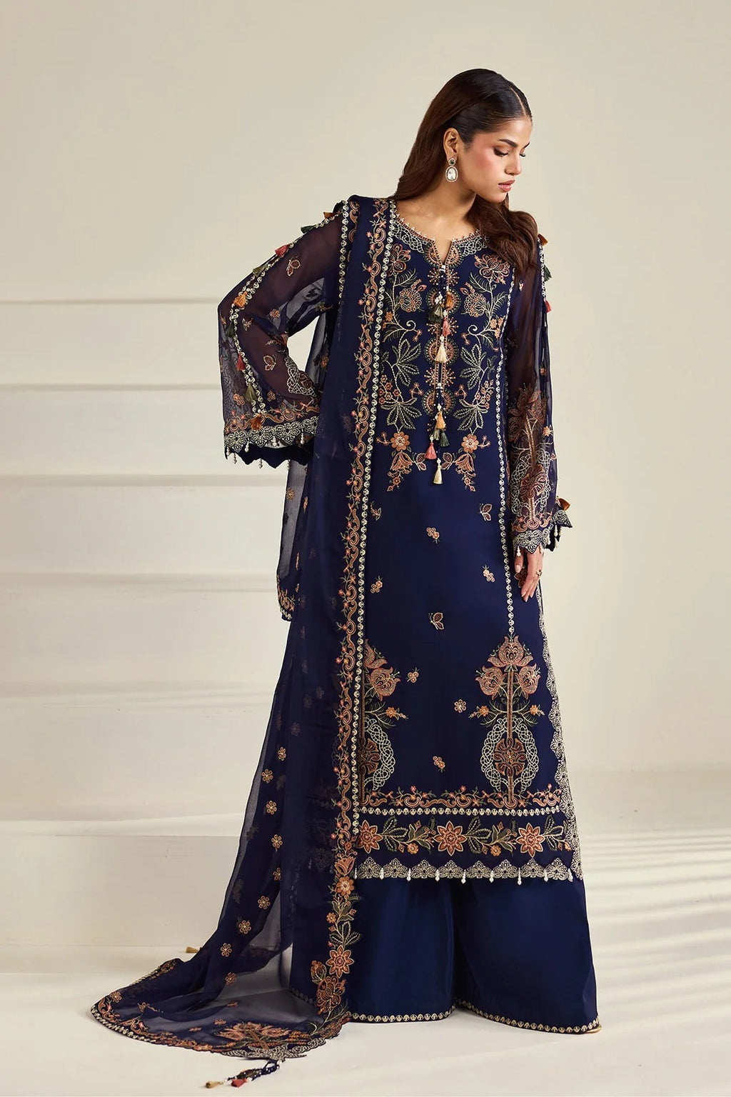 A woman models a traditional navy blue ethnic outfit with intricate gold and beige embroidery, featuring a long kurta, wide-leg pants, and a matching sheer dupatta, set against a neutral cream-colored background.