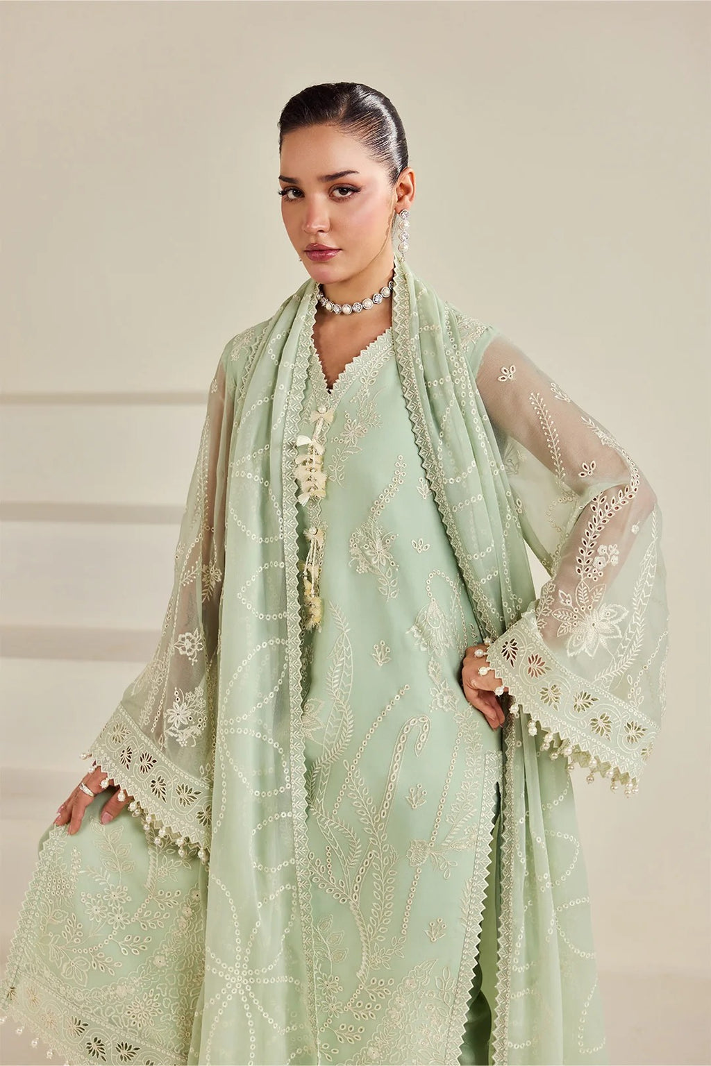 A woman models a traditional light green embroidered outfit with a matching sheer dupatta and white embroidered sleeves, set against a neutral beige background.