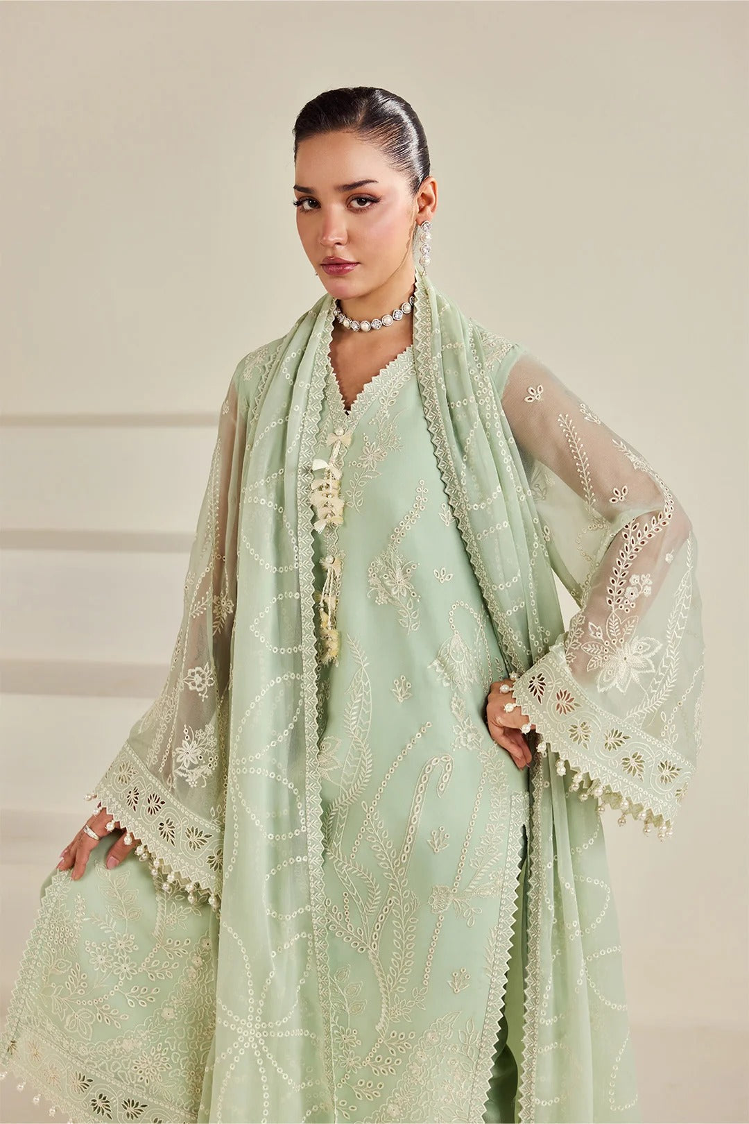A woman models a traditional light green embroidered outfit with a matching sheer dupatta and white embroidered sleeves, set against a neutral beige background.