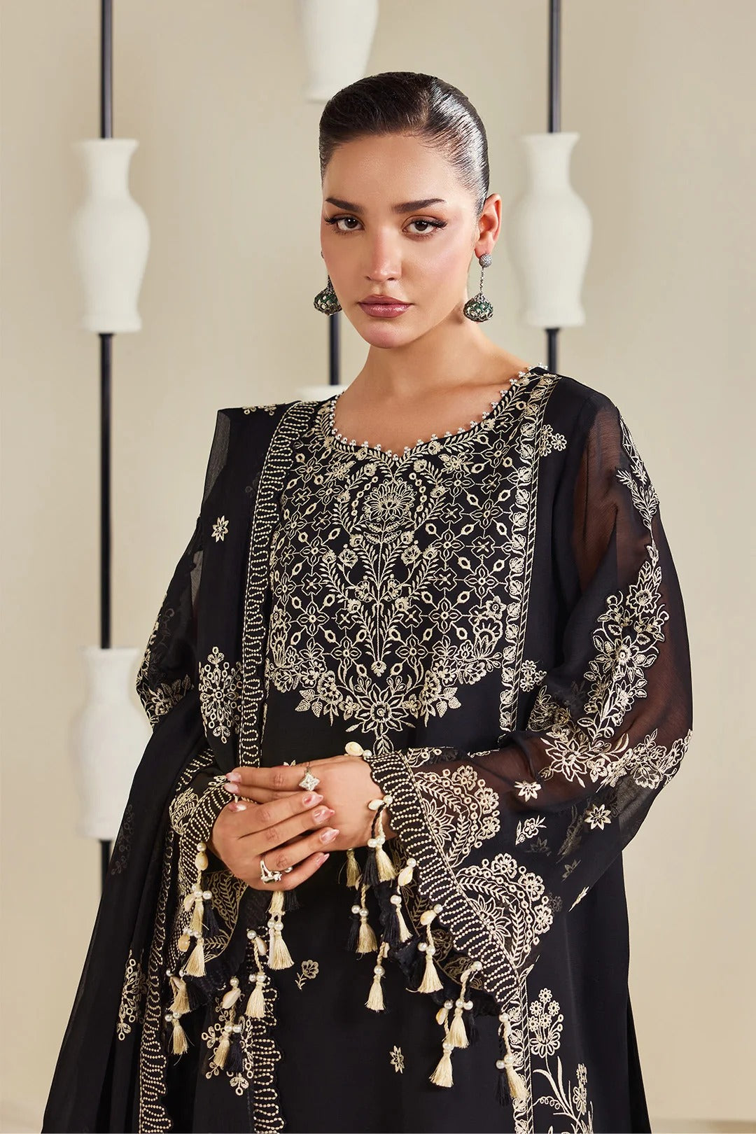 A woman modeling a black embroidered traditional outfit with intricate silver and gold detailing, sheer sleeves, and tassel accents, set against a neutral indoor background with decorative white vases.