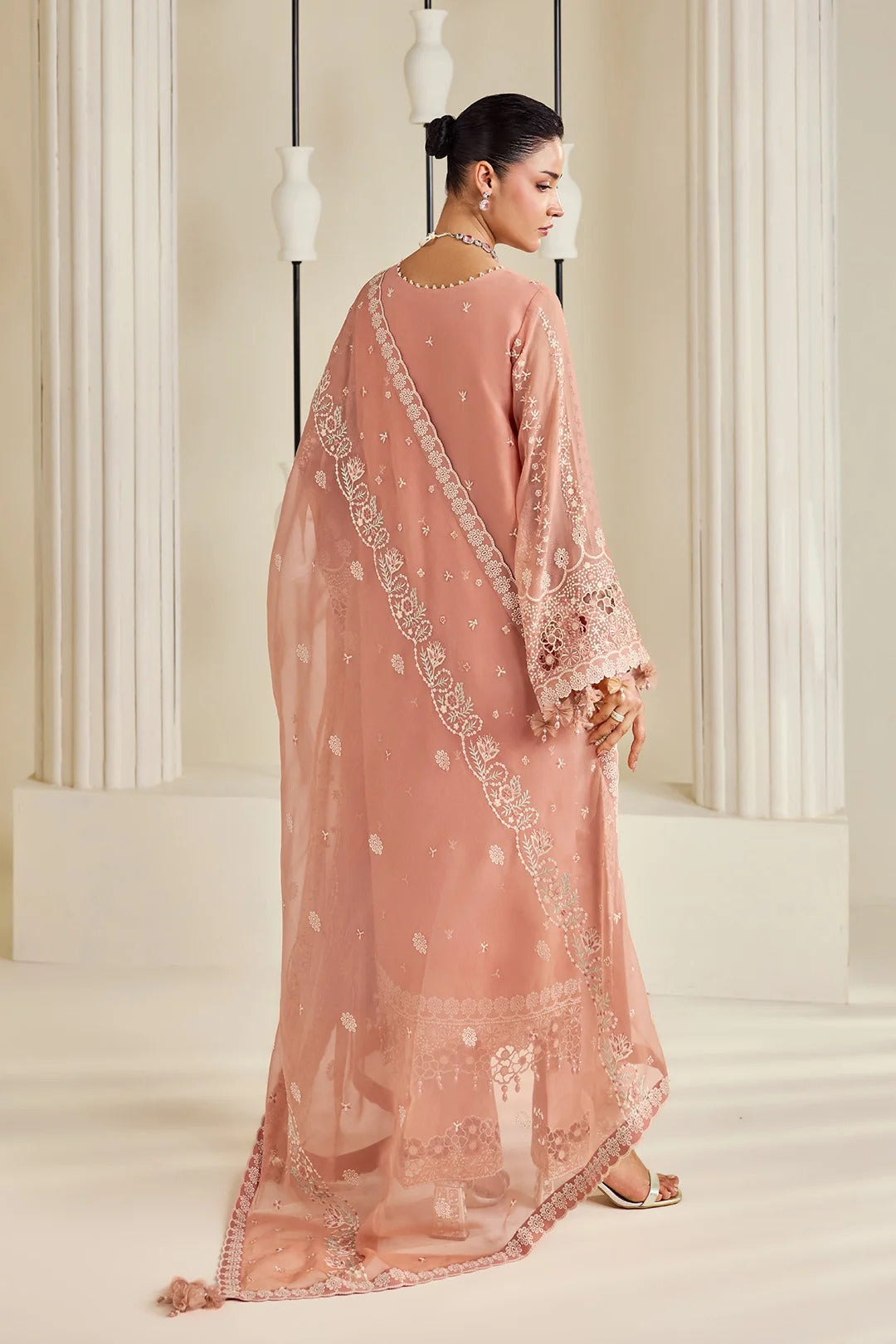 A woman modeling a peach-pink traditional outfit with intricate silver embroidery, shown from the back in a bright, elegant indoor setting with white columns.