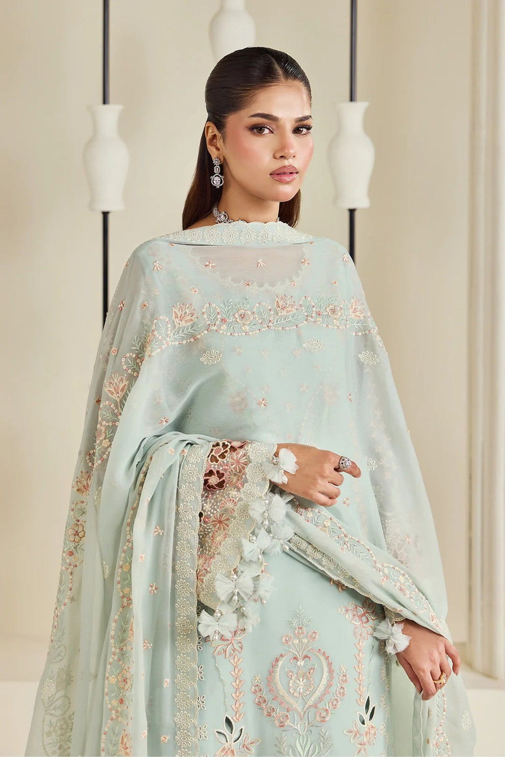 A woman modeling a light pastel mint green traditional outfit with intricate floral embroidery and a matching sheer dupatta, set against a warm beige background with decorative hanging vases.