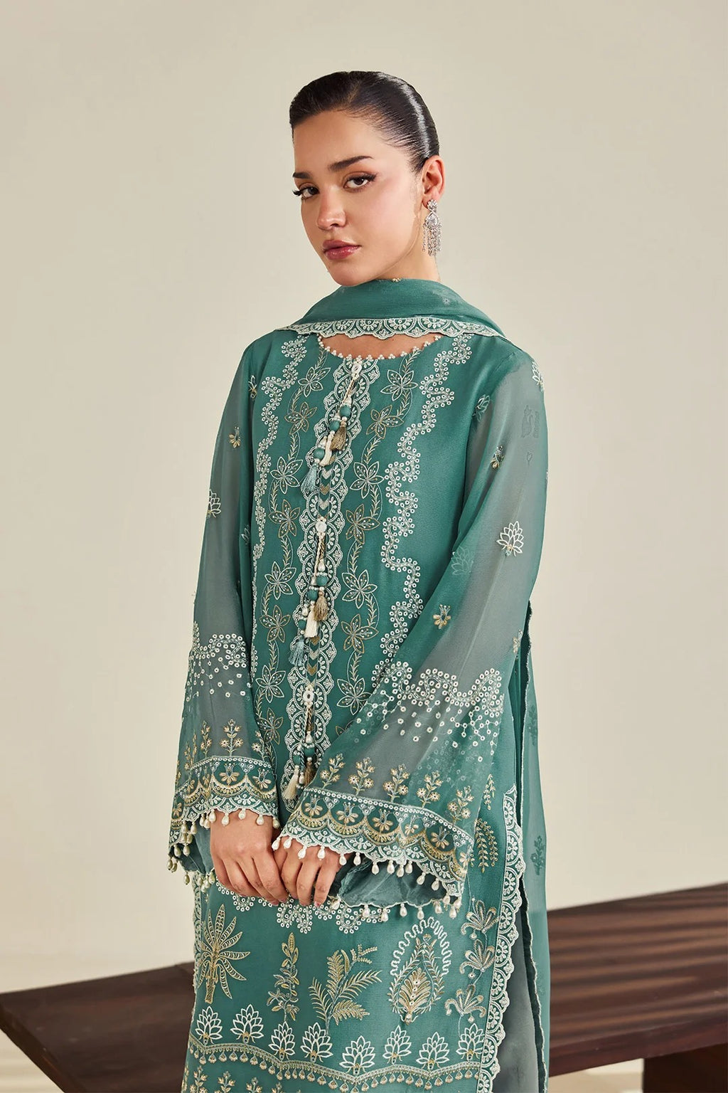 A woman modeling a teal green embroidered traditional outfit with a matching dupatta, featuring intricate gold and silver floral embroidery, set against a neutral beige background.