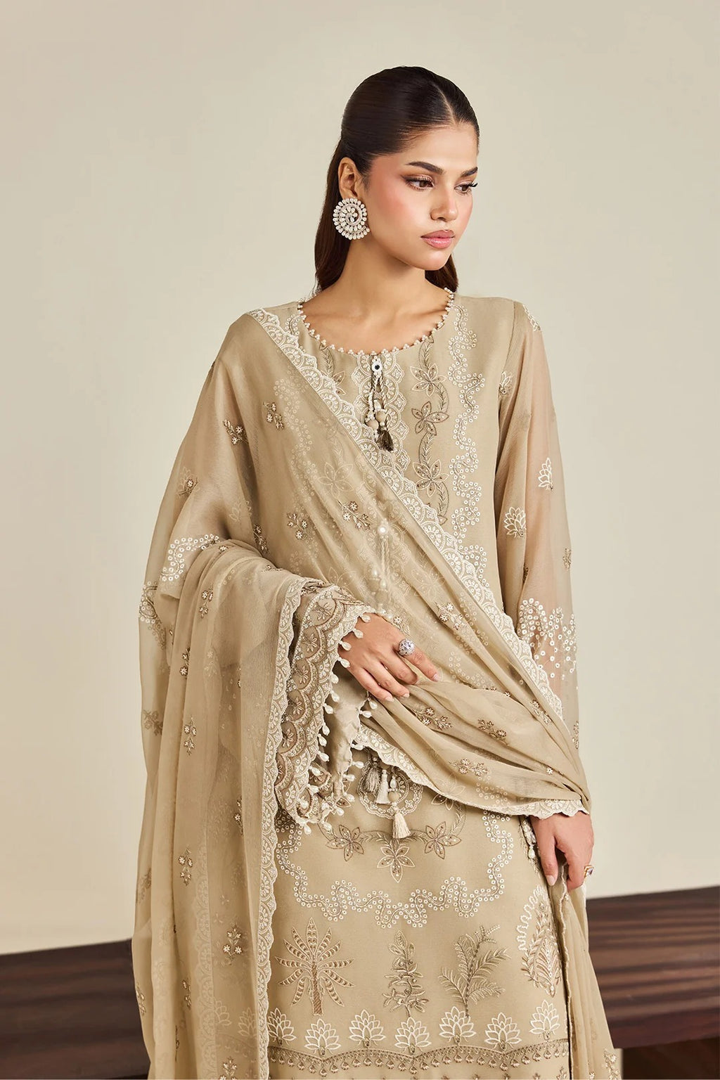 A woman models a beige traditional embroidered outfit with a matching dupatta, featuring intricate white floral embroidery and lace details, set against a neutral beige background.