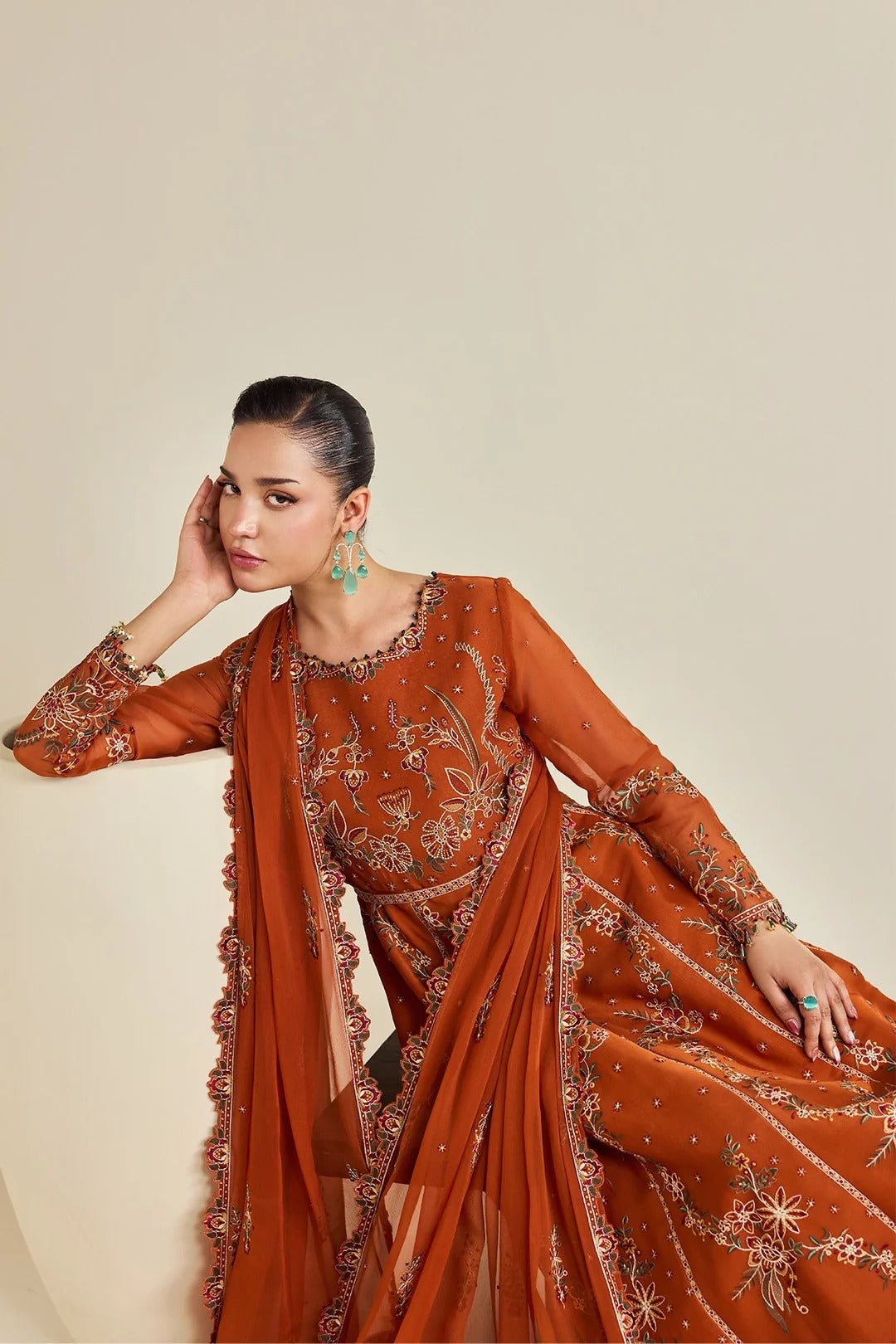 A woman models a rust-orange embroidered traditional outfit with a matching sheer dupatta, featuring intricate floral embroidery and gold detailing, posed against a solid beige background.