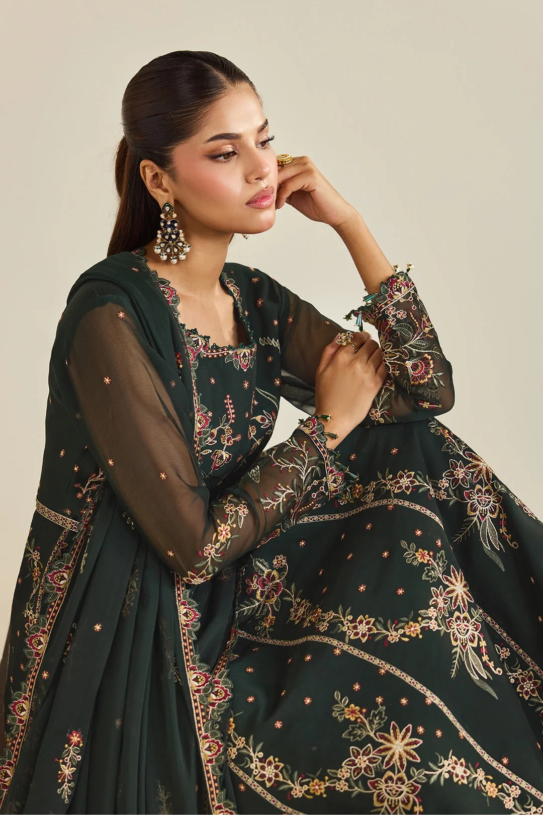 A woman models a dark green embroidered traditional outfit with sheer sleeves and a matching dupatta, set against a plain beige background.