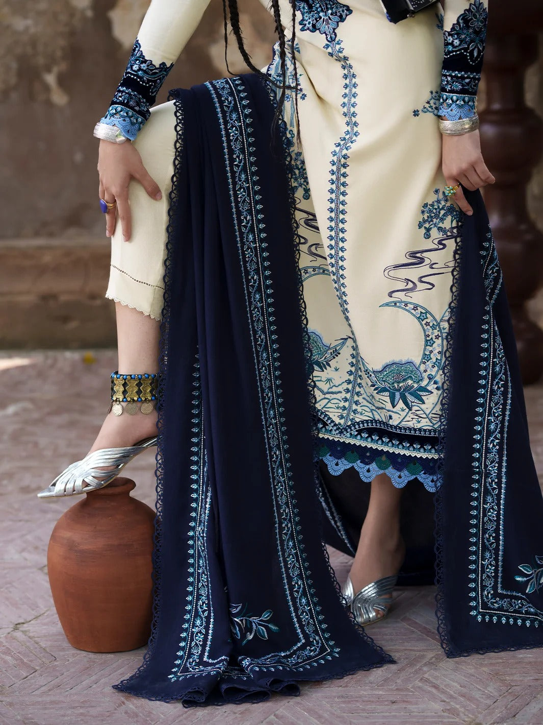 A model wearing a cream-colored embroidered kurta with a navy blue dupatta featuring intricate blue and white floral patterns, styled with silver shoes and jewelry in an outdoor setting.