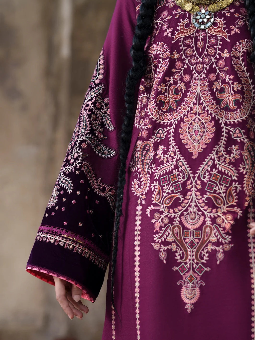 A close-up of a person wearing a rich magenta-purple traditional embroidered garment with intricate pink, gold, and white embroidery, featuring a long black braid and a decorative necklace.