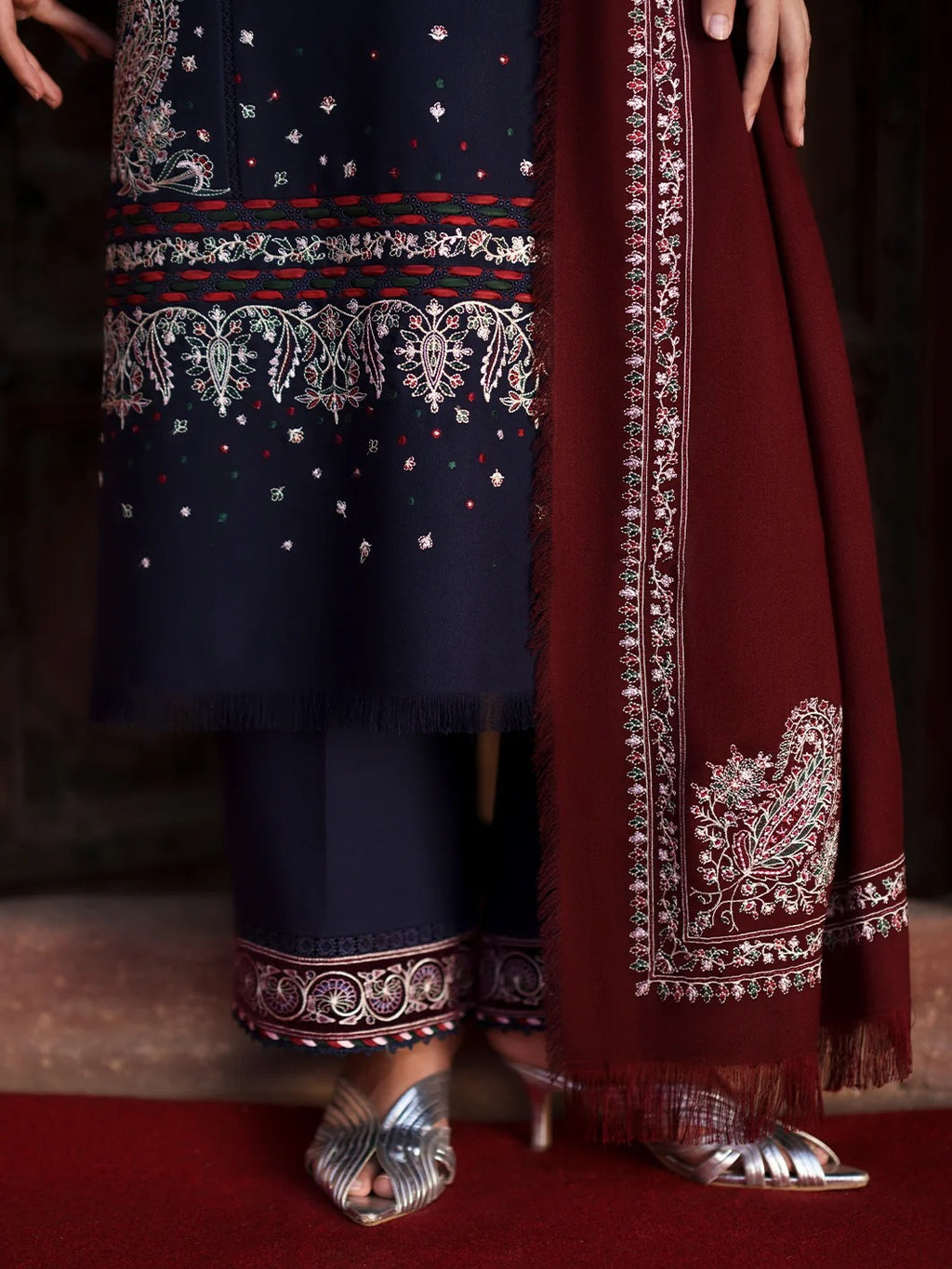 A close-up of a traditional South Asian outfit featuring a navy blue kurta with intricate silver and red embroidery, matching navy pants, and a deep maroon dupatta with ornate silver border detailing, styled with metallic silver sandals.