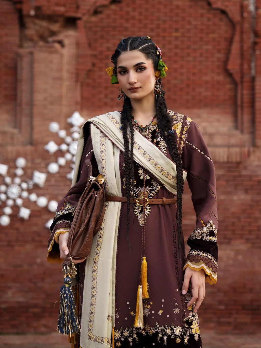 A woman wearing an ornate traditional outfit with a deep plum-brown tunic, cream draped scarf, and gold embroidery, standing in front of a red brick wall.