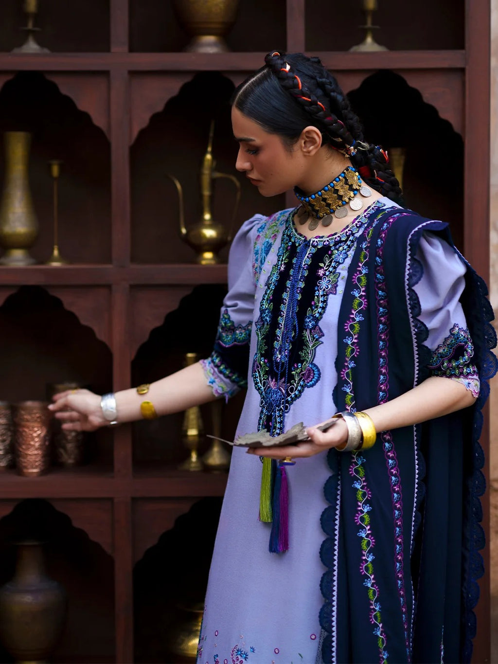 A woman wearing a traditional embroidered outfit with a lavender kurta, navy draped shawl, and ornate jewelry stands in front of a dark wooden shelving unit with decorative vases.