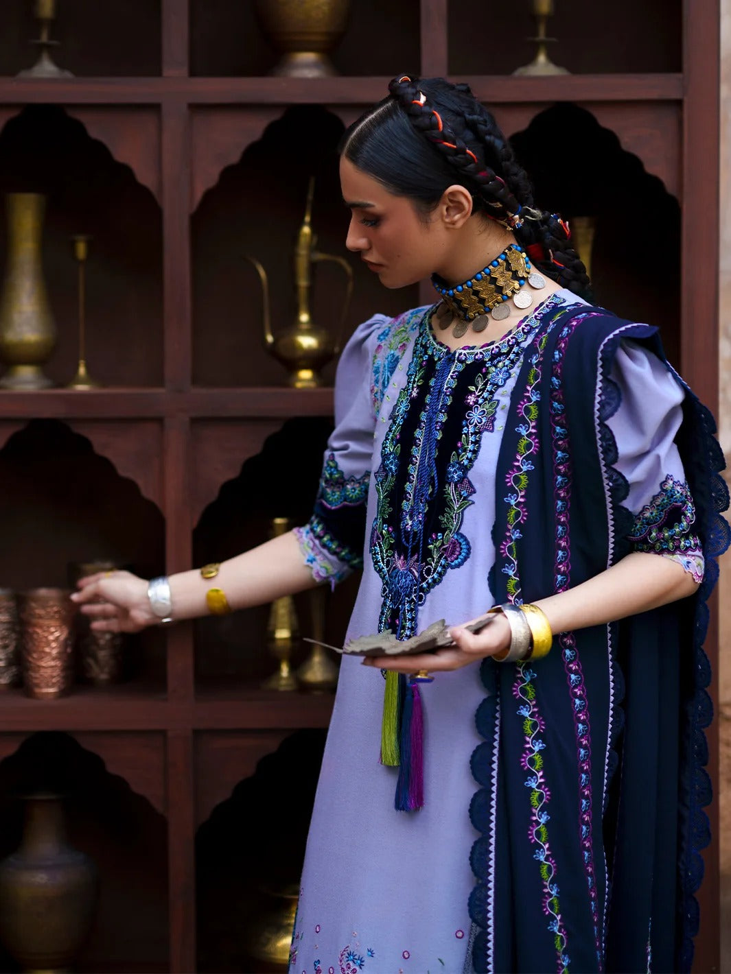 A woman wearing a traditional embroidered outfit with a lavender kurta, navy draped shawl, and ornate jewelry stands in front of a dark wooden shelving unit with decorative vases.