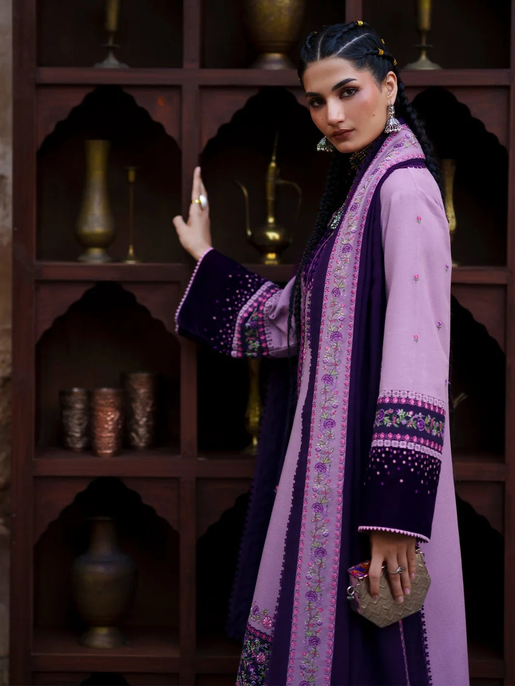 A woman models a traditional long-sleeve purple and lavender embroidered kurta with a matching dark purple shawl, standing in front of a dark wooden arched shelf with decorative brass vessels.