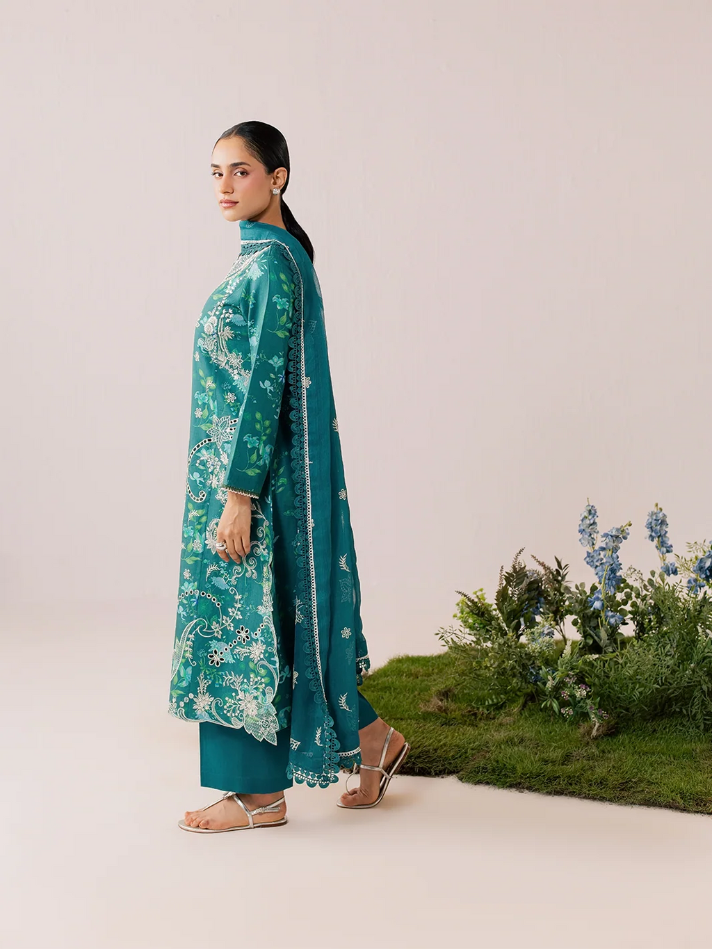 A woman modeling a teal green ethnic kurta set with white floral embroidery, matching pants, and a dupatta, standing in profile against a light beige studio backdrop with a small green plant arrangement at the bottom right.