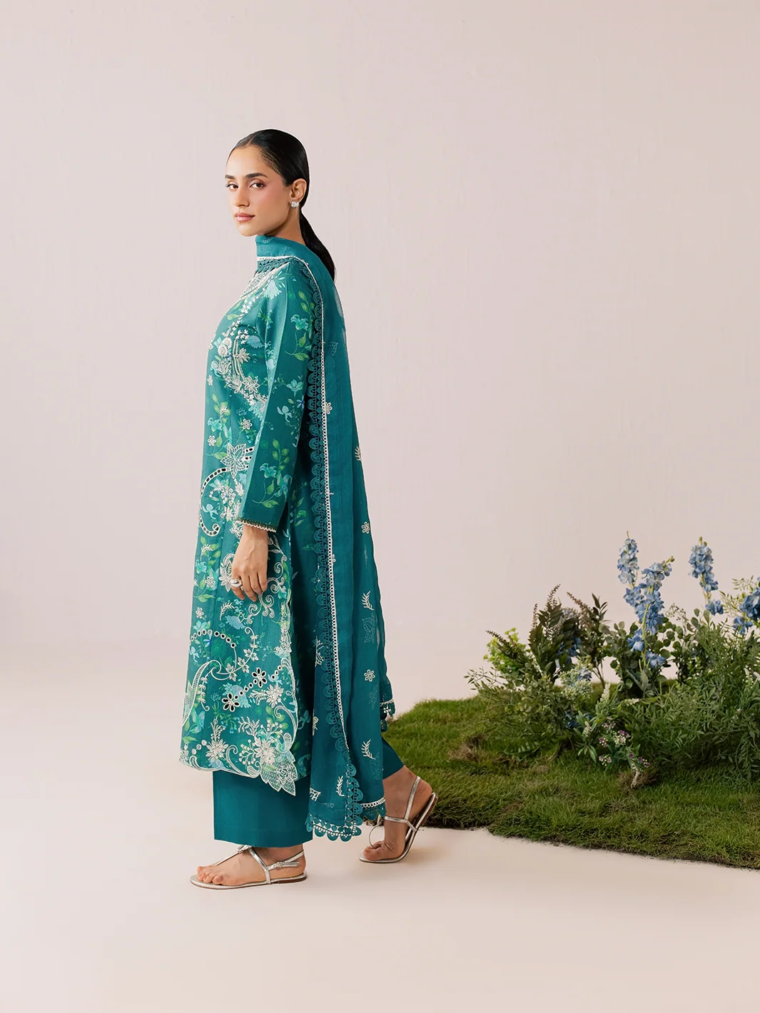 A woman modeling a teal green ethnic kurta set with white floral embroidery, matching pants, and a dupatta, standing in profile against a light beige studio backdrop with a small green plant arrangement at the bottom right.
