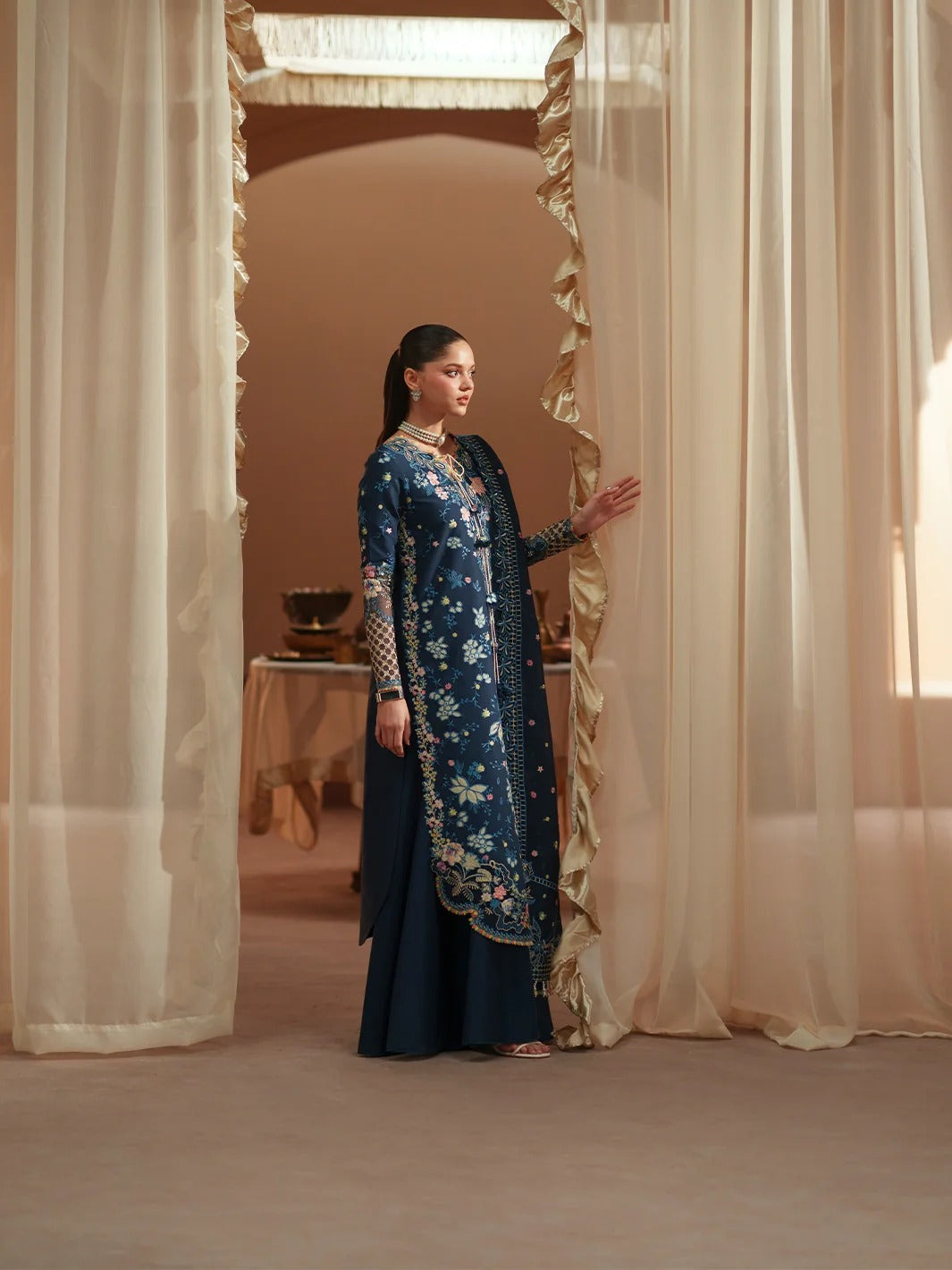 Woman in a blue floral dress standing between two curtains in a warm-toned room.