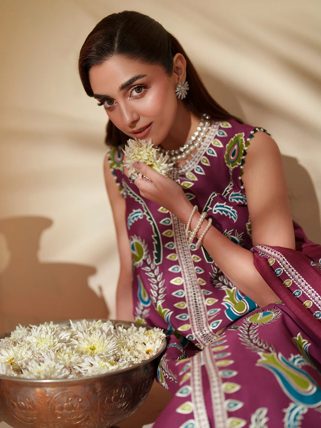 Woman in a colorful traditional outfit holding flowers and a bowl of flowers against a beige background