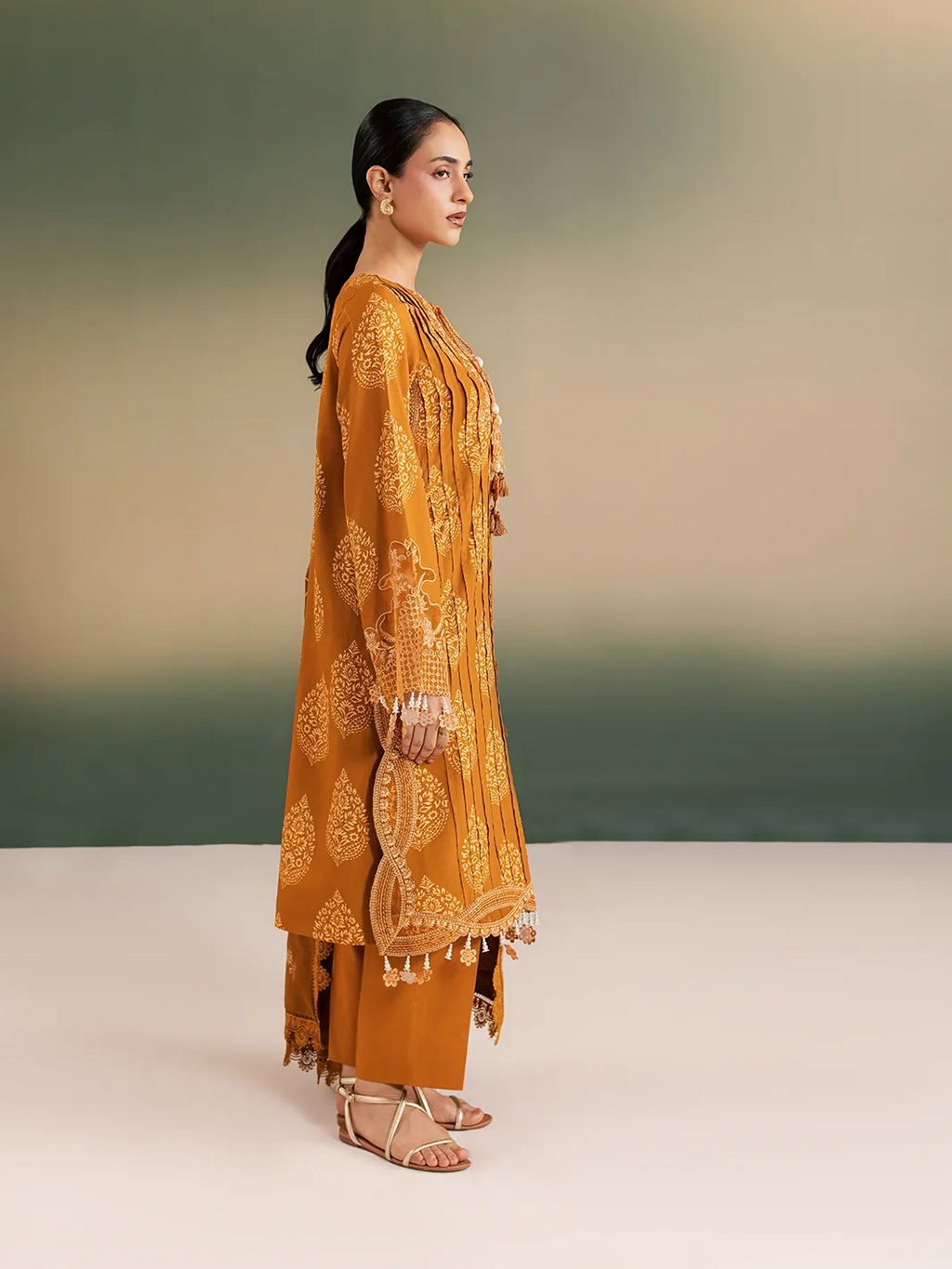 A woman modeling a mustard yellow traditional outfit with intricate gold embroidery and tassel details, standing in profile against a soft gradient studio background.