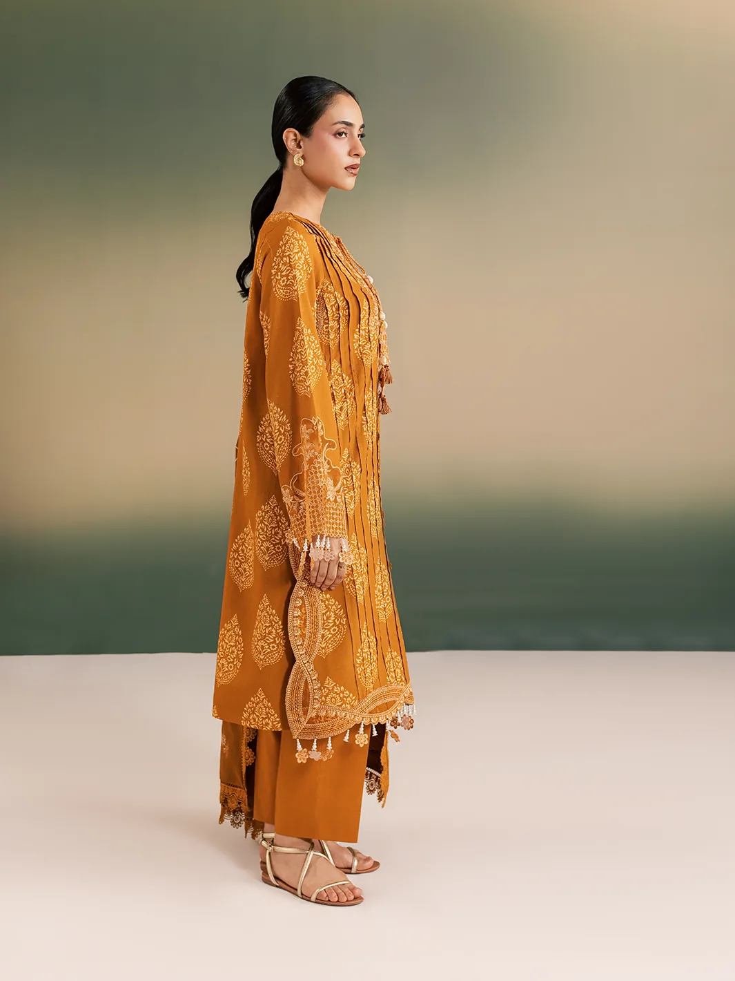 A woman modeling a mustard yellow traditional outfit with intricate gold embroidery and tassel details, standing in profile against a soft gradient studio background.