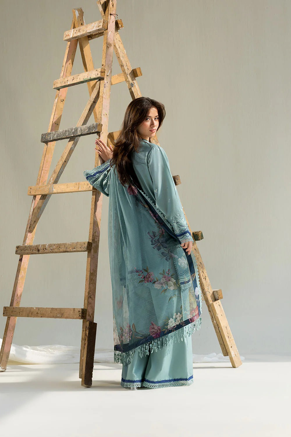 Woman in a light blue traditional outfit with floral patterns standing next to a wooden ladder.