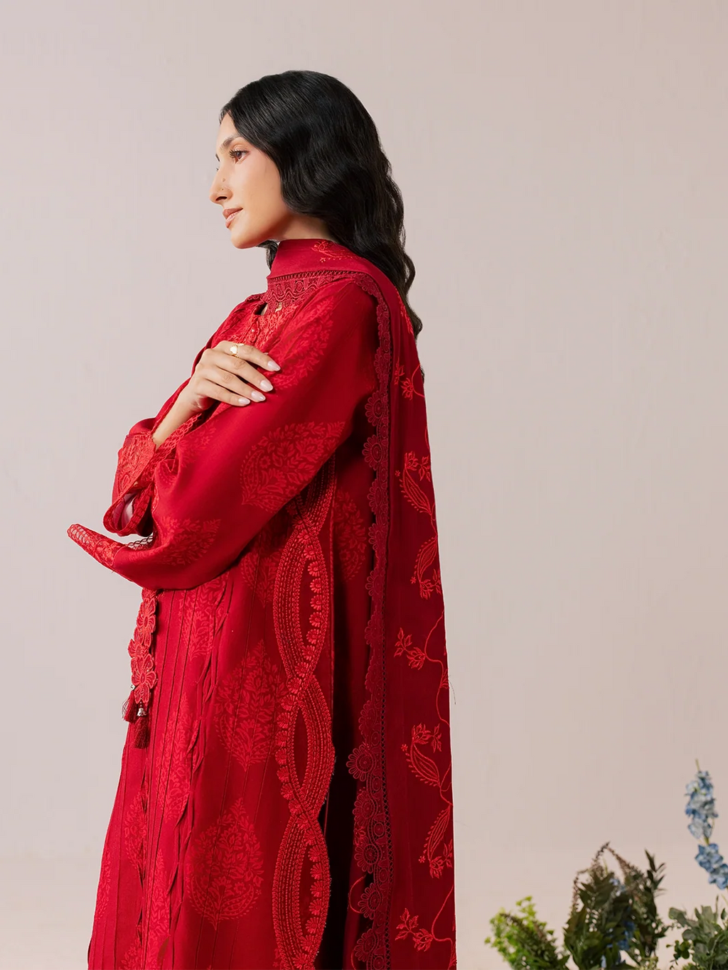 A woman in profile wearing a rich red embroidered traditional outfit with a matching draped shawl, set against a plain light beige background.