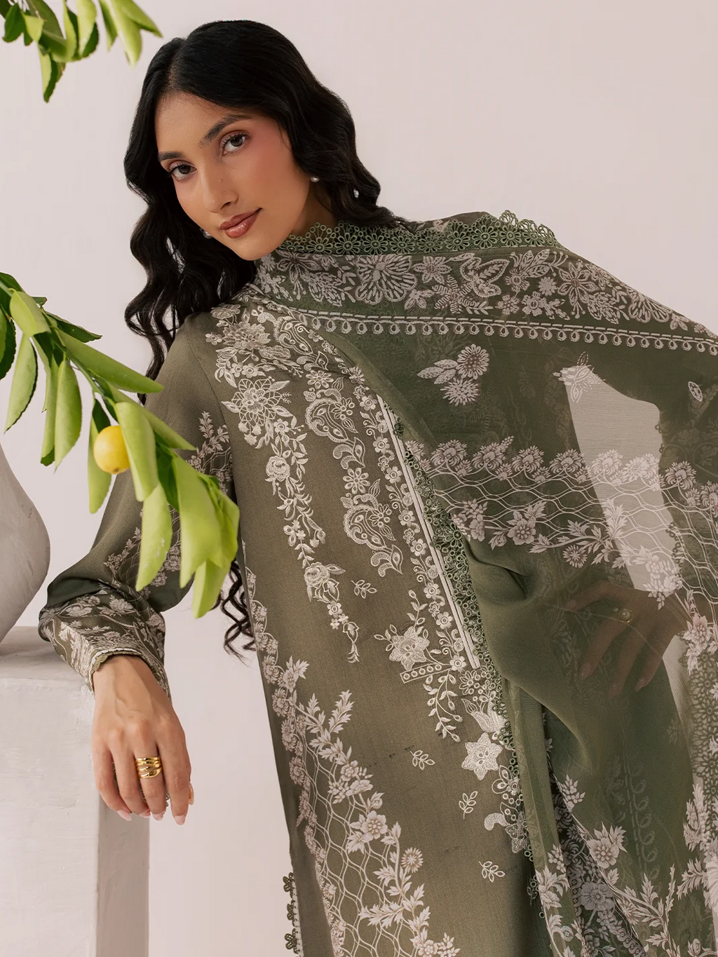 A woman models an olive green ethnic outfit with intricate white floral embroidery and a matching sheer dupatta, posed next to a white pillar with green leaves.