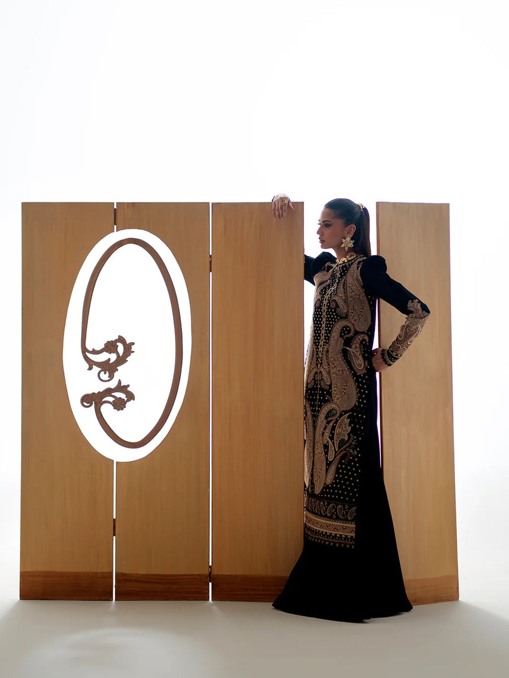 Woman in a long black dress standing next to a wooden screen with a logo on it.