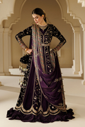 A woman models a traditional Indian ethnic outfit consisting of a deep purple velvet lehenga, a heavily embroidered black kurta, and a matching draped dupatta, set against an elegant beige stone archway background.