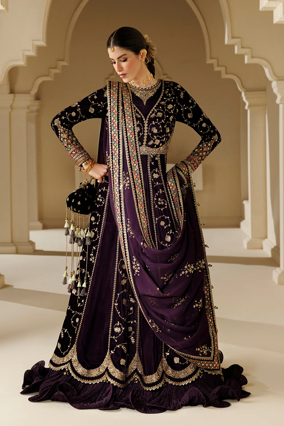 A woman models a traditional Indian ethnic outfit consisting of a deep purple velvet lehenga, a heavily embroidered black kurta, and a matching draped dupatta, set against an elegant beige stone archway background.