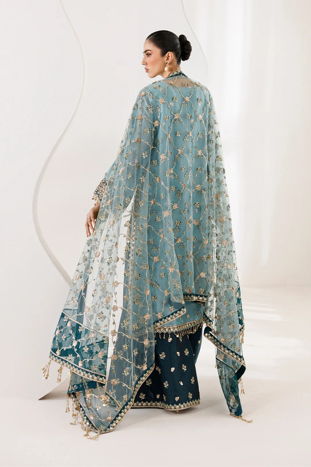 A woman modeling a traditional South Asian outfit with a teal blue embroidered dupatta draped over her shoulders, paired with dark navy flared pants featuring gold borders, photographed in a bright studio setting.