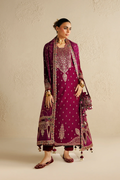 A woman models a traditional South Asian outfit consisting of a deep magenta kurta with gold embroidery, matching trousers, a matching dupatta, and flat shoes, set against a textured beige wall.