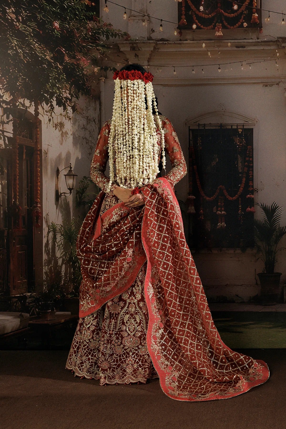 A woman photographed from behind wearing an ornate traditional bridal outfit with a long cream floral veil, red and gold embroidered skirt, and a draped red patterned dupatta, standing in a decorated courtyard.