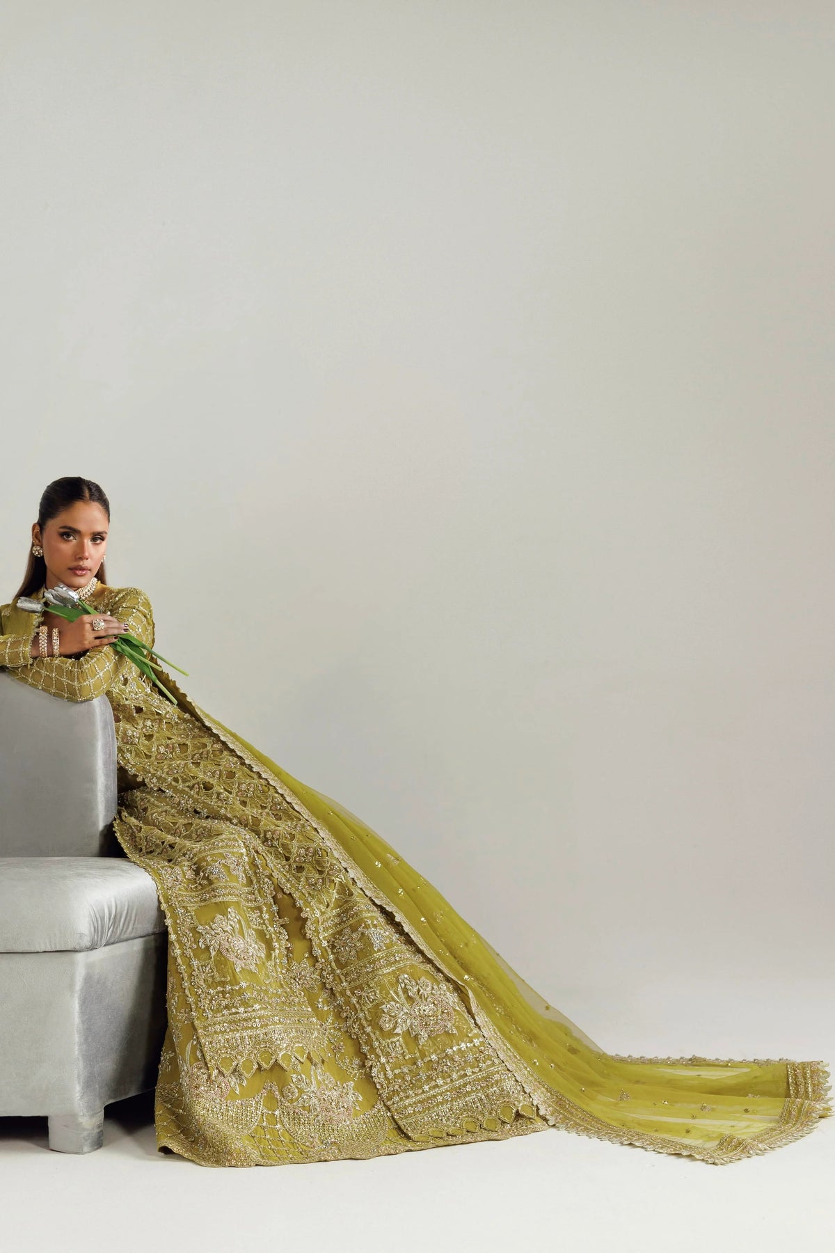 A model wearing a heavily embroidered olive green traditional gown with a long train, seated on a gray upholstered chair against a plain light gray studio background.