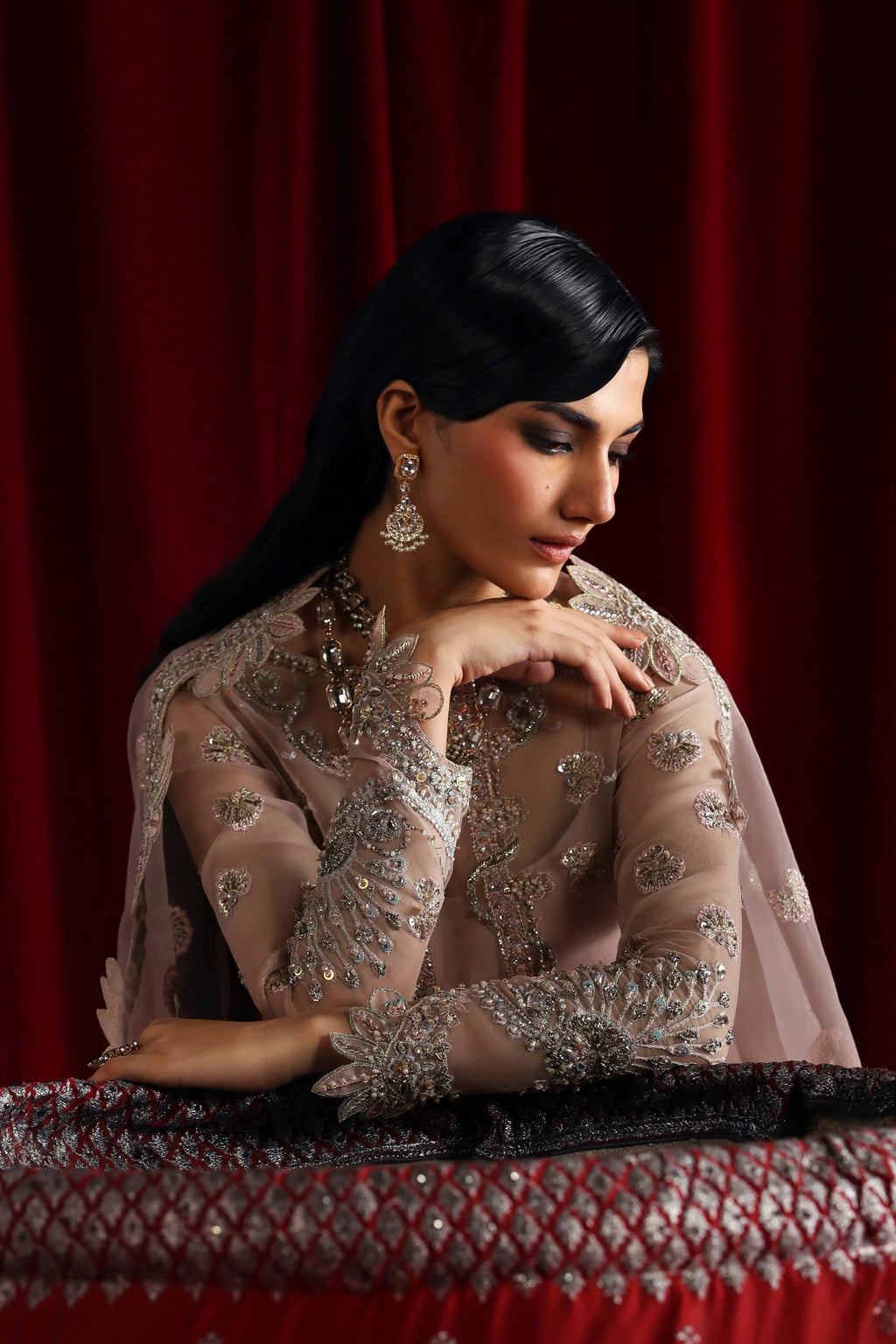 A woman wearing an ornate beige embroidered traditional outfit with sheer sleeves and heavy silver beadwork, posed against a deep red curtain backdrop.