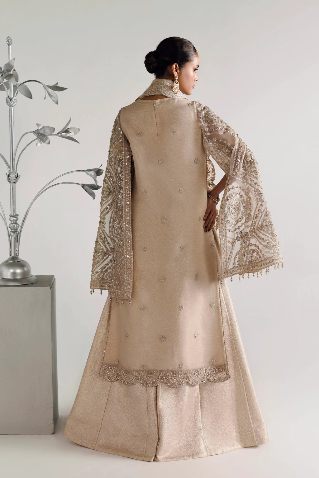 A woman shown from the back wearing a beige traditional outfit with ornate silver embroidery and a matching embroidered shawl, photographed against a light gray studio background with a decorative silver plant on the left.