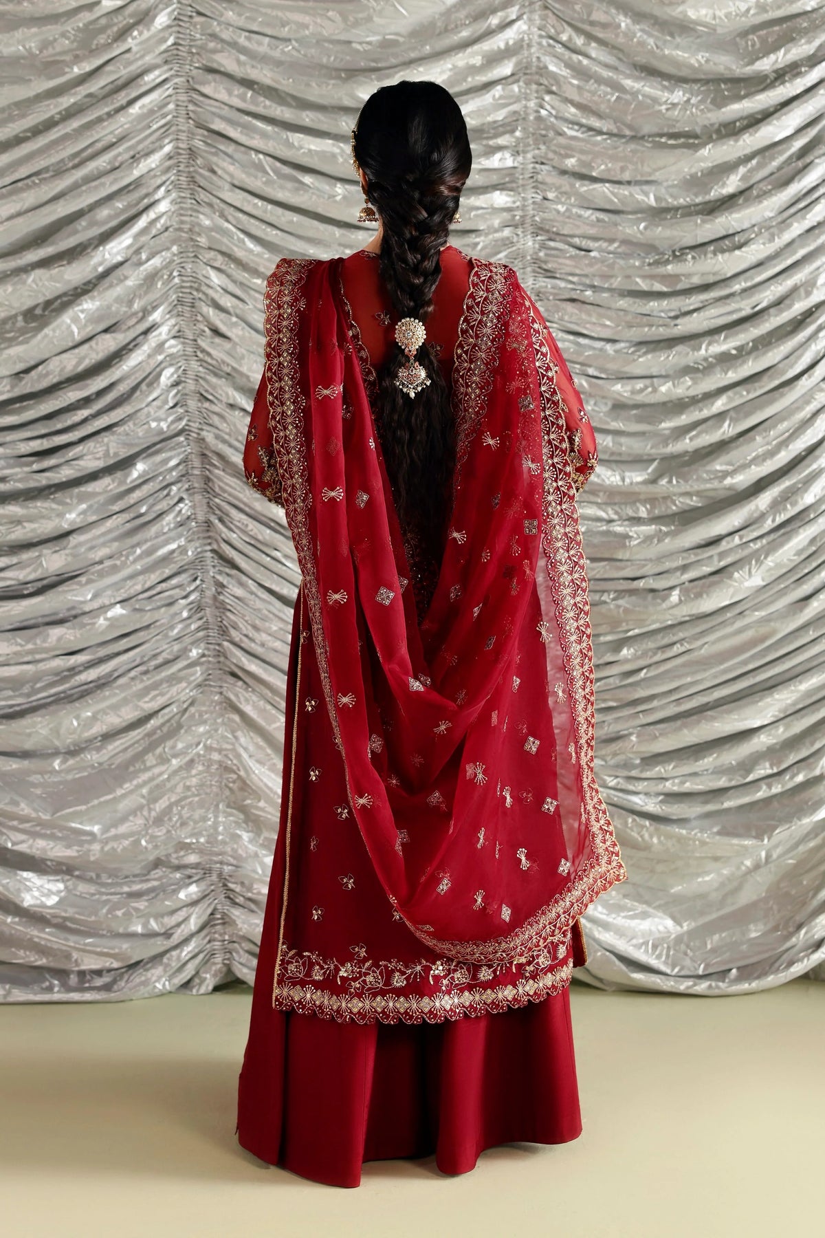 A woman shown from the back wearing a traditional red outfit with a matching sheer dupatta, both decorated with intricate gold and silver embroidery. She stands in front of a silver pleated backdrop.