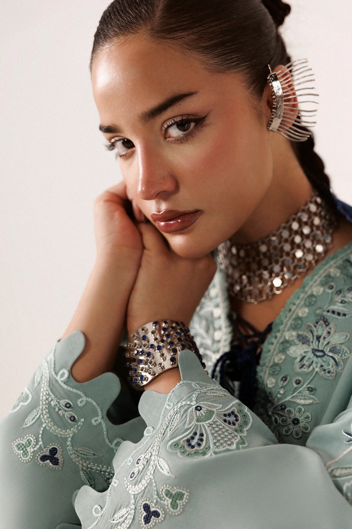 A close-up portrait of a woman wearing ornate silver jewelry, including a chunky choker necklace, a wide cuff bracelet, and a large hair clip, paired with a light blue embroidered traditional garment.