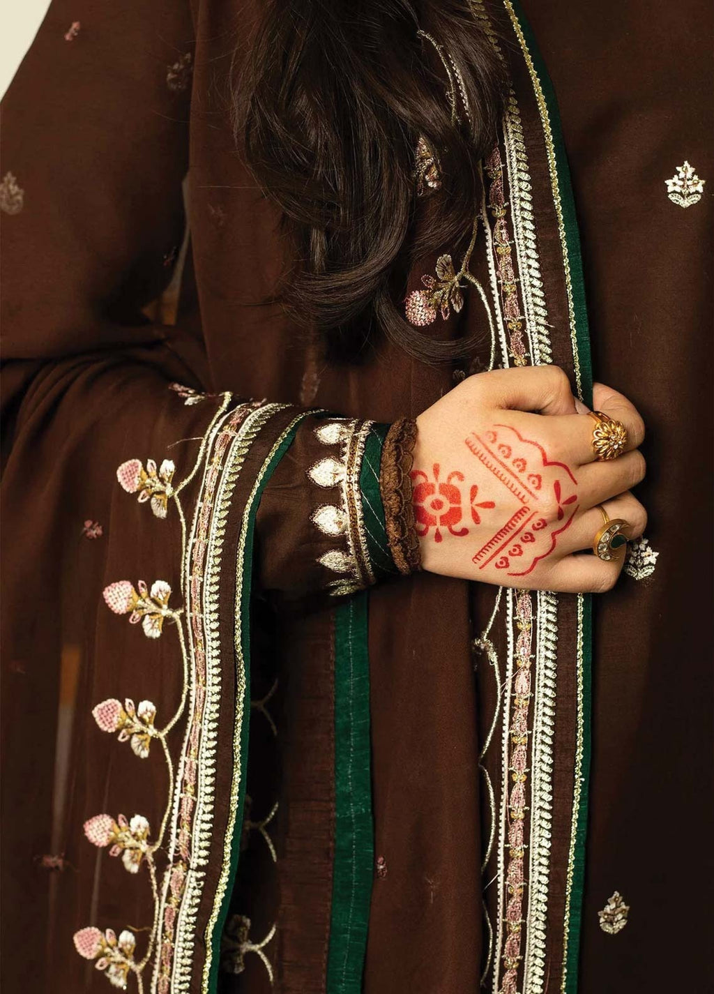 A close-up of a person wearing a dark brown traditional outfit with ornate silver and green embroidered borders, holding a matching brown dupatta. The person's hand features red henna designs and is adorned with gold rings.